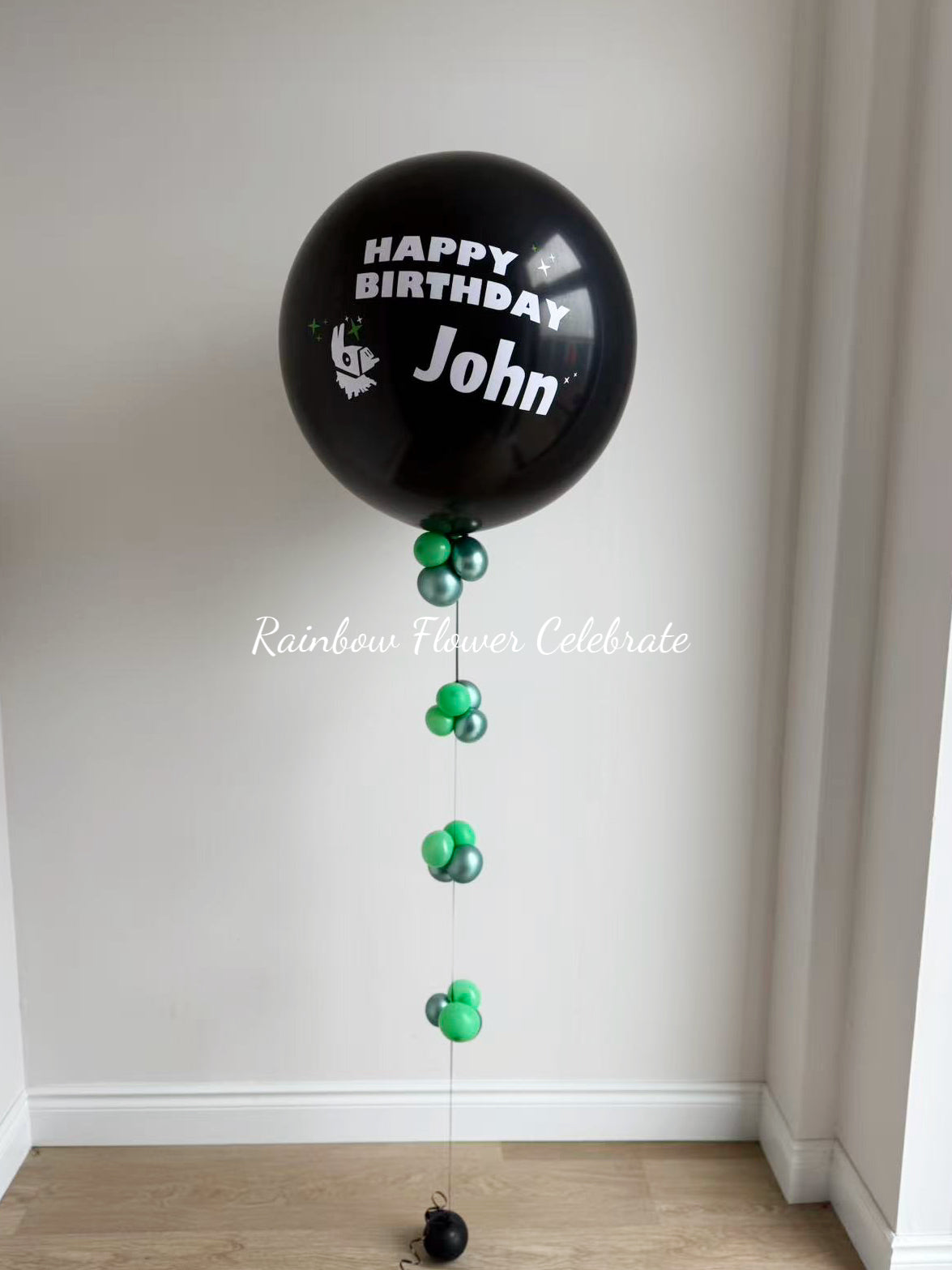 Black latex balloon with personalized message 'Happy Birthday John' with green mini balloons on string.