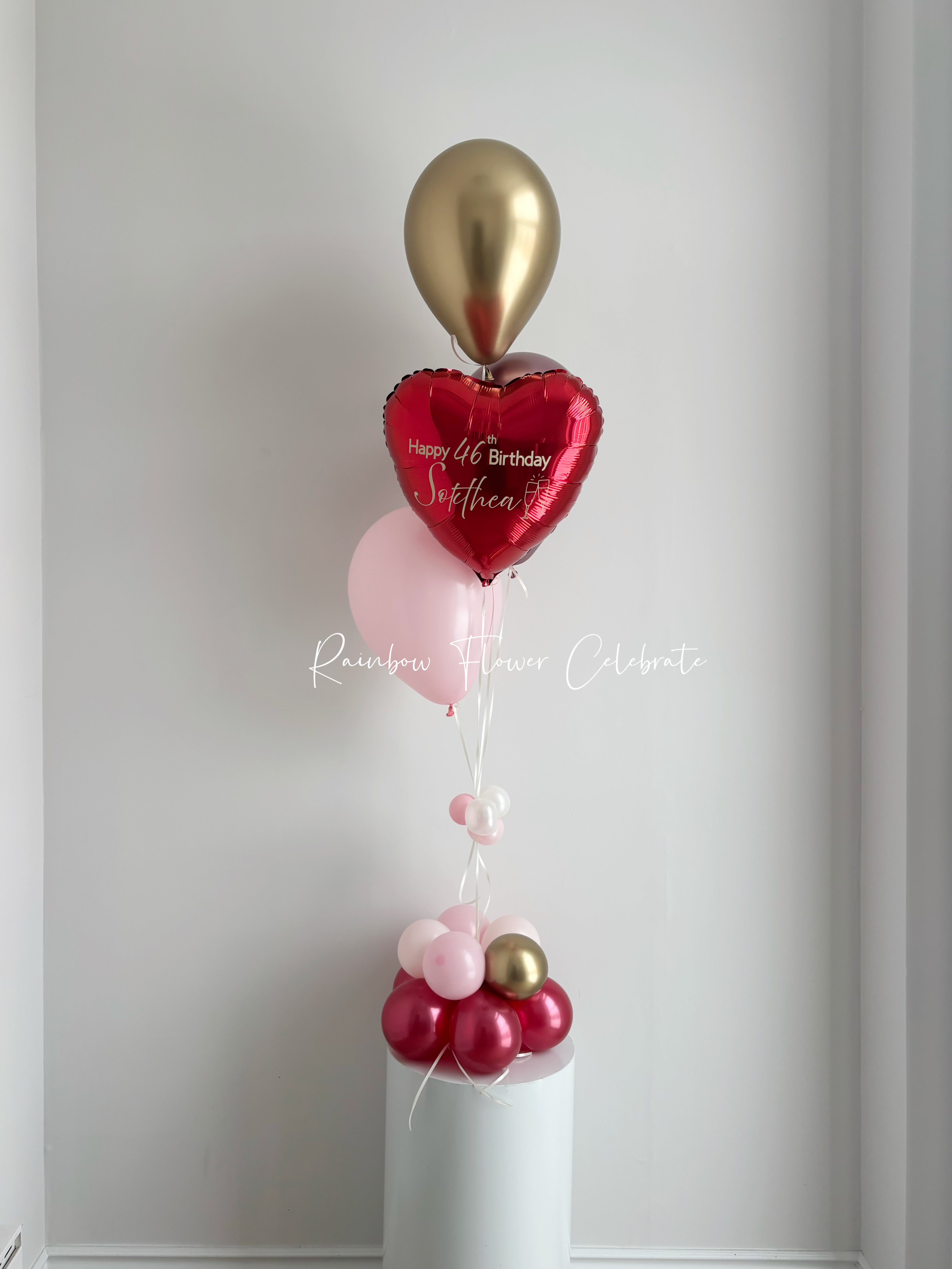 Birthday balloon bouquet with red heart, gold, and pink balloons on white pedestal