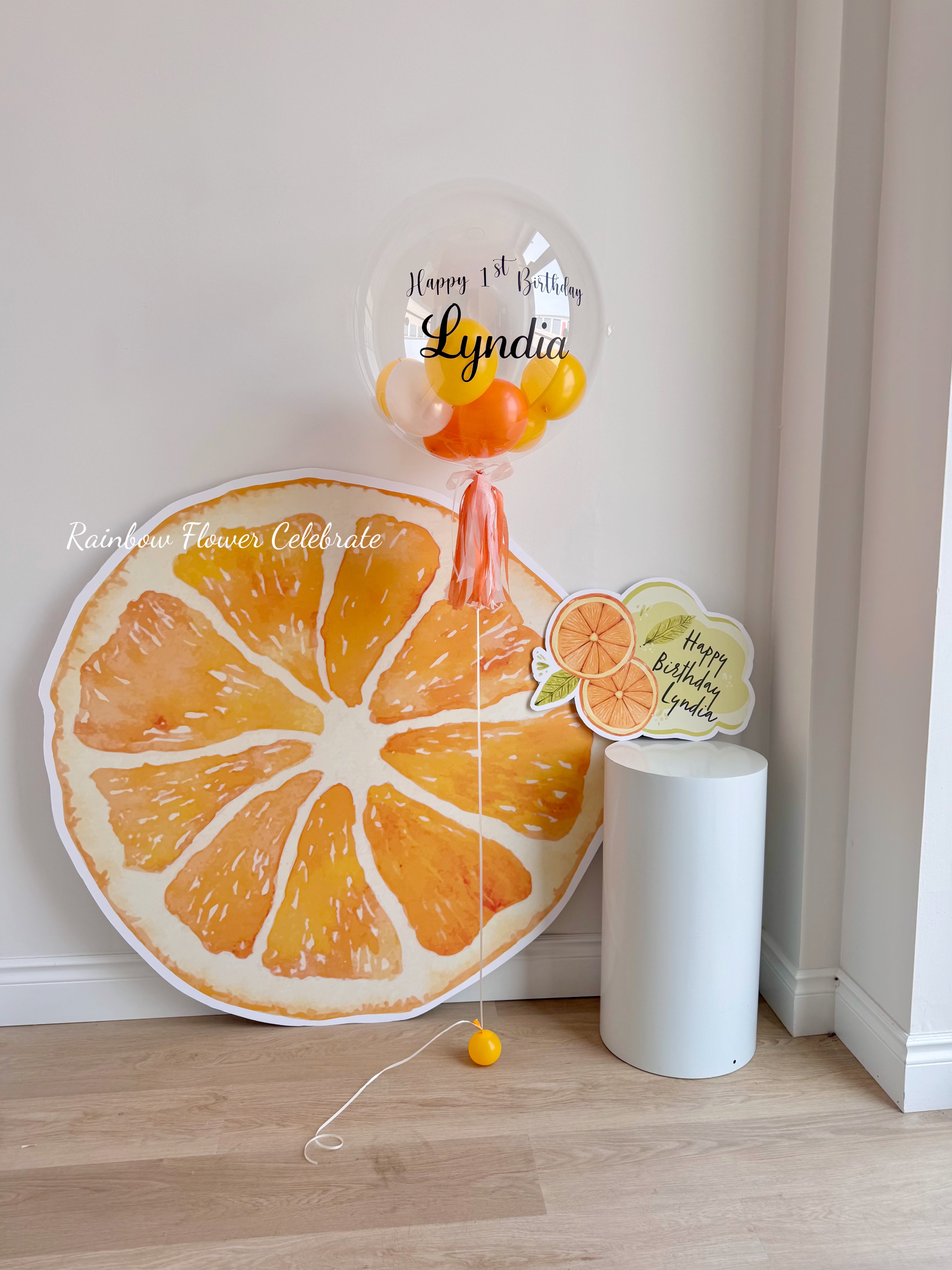 Decorative balloons shaped like an orange and a bubble with 'Happy 1st Birthday Lyndia' text on a white background.