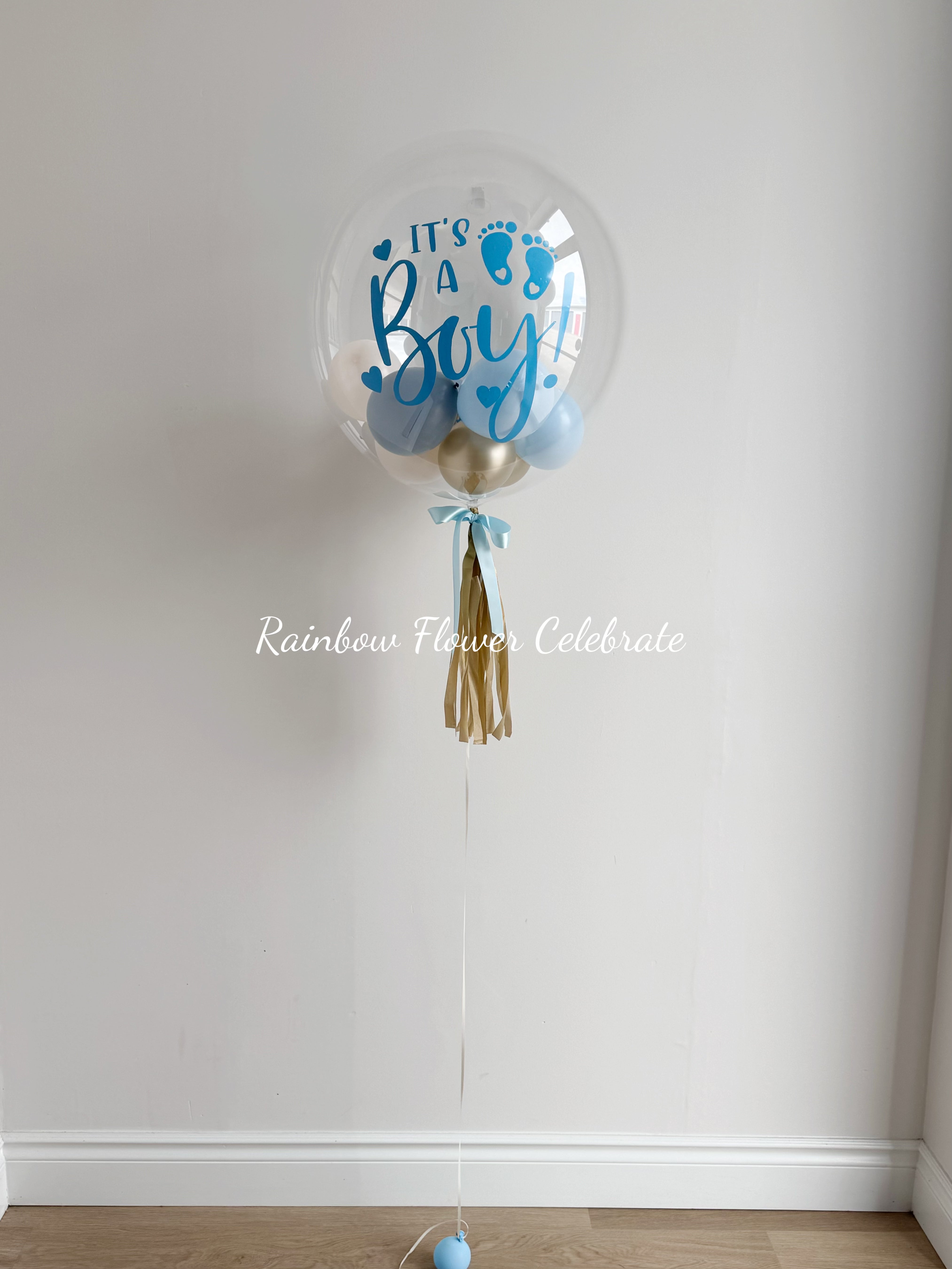 Clear balloon with 'It's a Boy' text and tassel against a white wall, featuring 'Rainbow Flower Celebrate' branding.